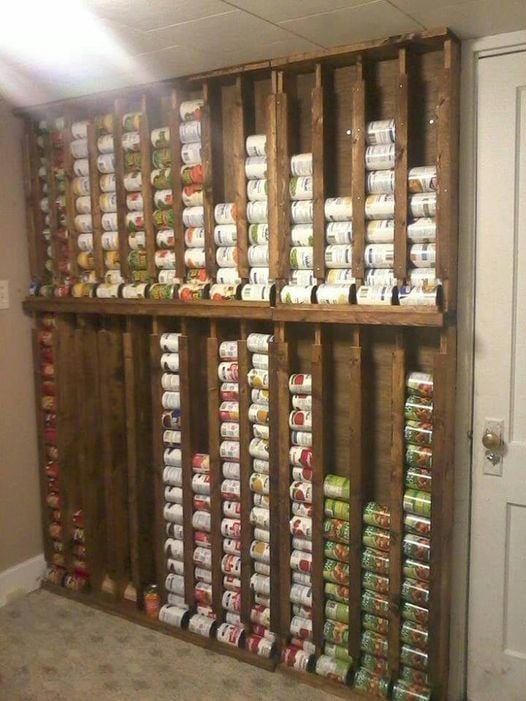 The meme is a photo of a wall with shelves that have been converted into a storage space for canned goods. The shelves are filled to the brim with cans of food, with some even spilling over onto the floor. The text "I'm not hoarding, I'm just stockpiling" appears at the top of the image.